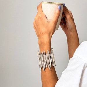 Könnte beinhalten: Eine Person in einem weißen Hemd und einem silbernen Armband mit einem einzigartigen Design hält eine weiße Keramiktasse mit einem hellbraunen Rand. Das Armband besteht aus vielen kleinen, dünnen, silbernen Teilen, die in einem vertikalen Muster angeordnet sind.