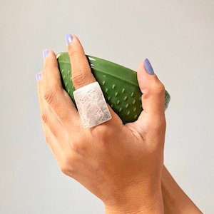 Könnte beinhalten: Ein silberner Ring mit einem strukturierten rechteckigen Design. Der Ring wird am Zeigefinger einer Hand getragen, die eine grüne Keramikschale hält.