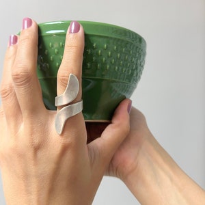 Könnte beinhalten: Ein silberner Ring mit einem einzigartigen, abstrakten Design. Der Ring wird am Zeigefinger einer Hand getragen, die eine grüne Keramikschale hält.