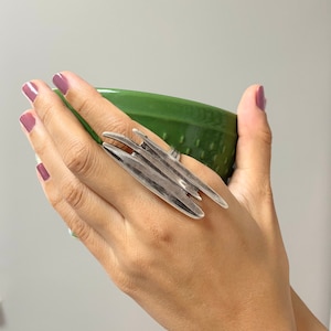 Könnte beinhalten: Ein silberner Ring mit geometrischem Design. Der Ring besteht aus mehreren dünnen, flachen Metallteilen, die in einem unregelmäßigen, abstrakten Muster angeordnet sind.
