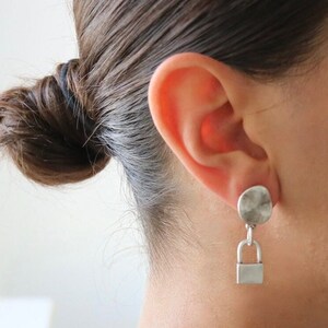 May include: Silver lock and key dangle earrings. The earrings feature a round silver disc with a textured surface and a silver lock charm hanging from it.