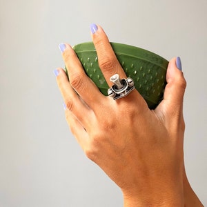 Könnte beinhalten: Ein silberner Ring mit einem einzigartigen Design, das eine kleine, erhabene, trompetenartige Form und dekorative Details aufweist. Der Ring wird an einer Hand getragen, die eine grüne Schüssel hält.