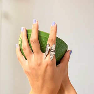 May include: A silver ring with two angel wings, worn on a finger holding a green bowl.