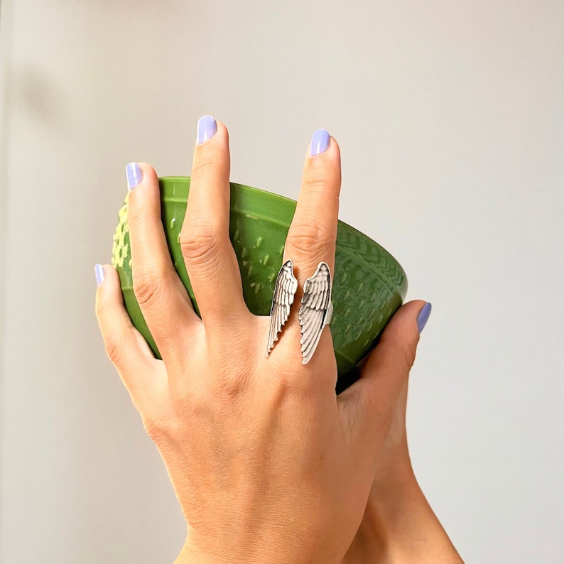 May include: A silver ring with two angel wings, worn on a finger holding a green bowl.