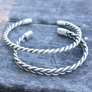 Könnte beinhalten: Zwei silberne geflochtene Armreifen. Die Armreifen sind aus gedrehtem Metall und haben ein schlichtes, elegantes Design.