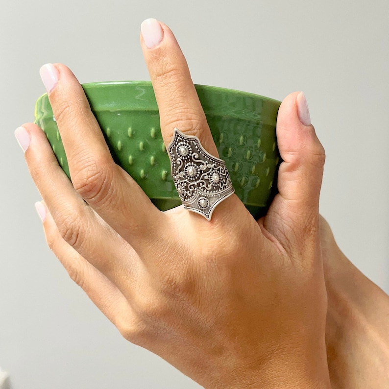 May include: A silver ring with an intricate design featuring a pointed top and floral details. The ring is worn on the index finger of a hand holding a green bowl.