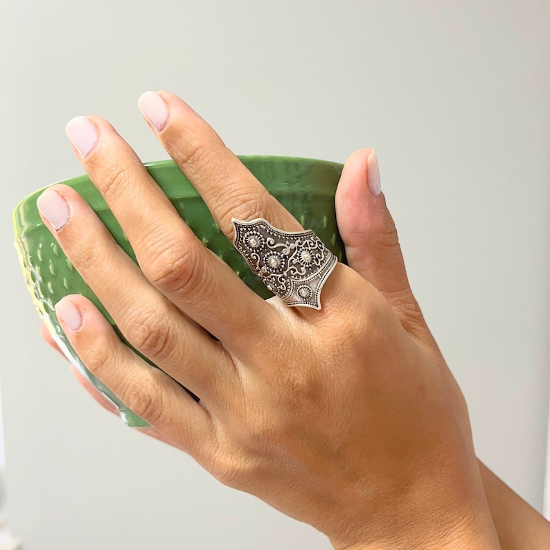 May include: A silver ring with an intricate design featuring a floral pattern. The ring is worn on the index finger of a hand holding a green bowl.