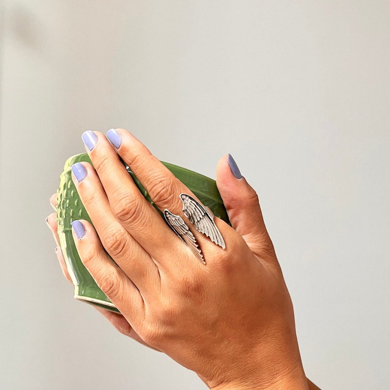 May include: A silver ring with a wing design. The ring is worn on the index finger of a hand holding a green ceramic mug.