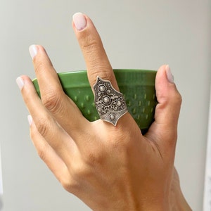 May include: A silver ring with an intricate design featuring a pointed shape and small round accents. The ring is worn on a finger holding a green bowl.