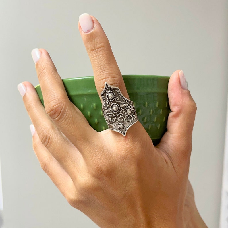 May include: A silver ring with an intricate design featuring a pointed shape and small round accents. The ring is worn on a finger holding a green bowl.