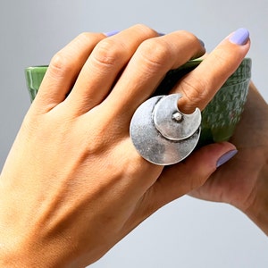 May include: A silver ring with a unique design featuring two overlapping circles. The ring is worn on a finger and is a statement piece.