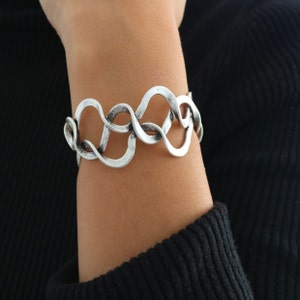 Könnte beinhalten: Ein silbernes Armband mit einem einzigartigen, abstrakten Design. Das Armband besteht aus gedrehtem Metall und hat eine wellige, organische Form.