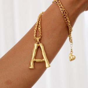 Könnte beinhalten: Eine goldene Kettenkette mit einem dicken, strukturierten Buchstaben 'A' als Anhänger. Die Kette ist mit einer kleineren, filigraneren Kette mit einem Herz-Anhänger geschichtet.