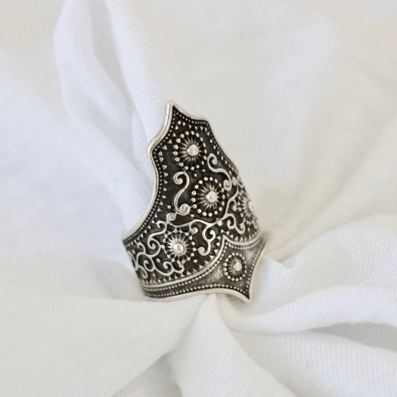 May include: A silver ring with an intricate floral design. The ring is wide and has a pointed top.