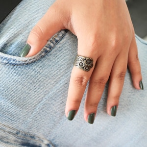 Könnte beinhalten: Ein silberner Ring mit floralem Design. Der Ring wird am Zeigefinger einer Hand mit grünem Nagellack getragen.