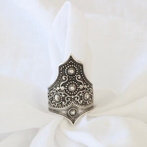 May include: A silver ring with an intricate, ornate design. The ring is shaped like a crown with a pointed top and a wide band.