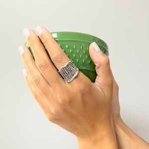 May include: A silver ring with a textured, organic design. The ring is wide and has a slightly curved shape.