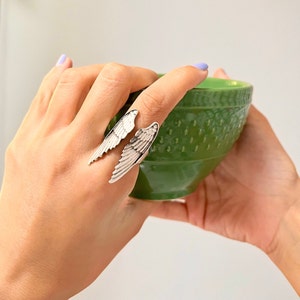 May include: A silver ring with two wings that wrap around the finger. The ring is on a person's finger, and the person is holding a green bowl.