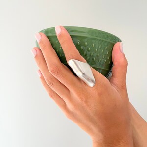 Könnte beinhalten: Ein silberner Ring mit einem einzigartigen, eckigen Design. Der Ring wird am Zeigefinger einer Hand getragen, die eine grüne Schüssel hält.