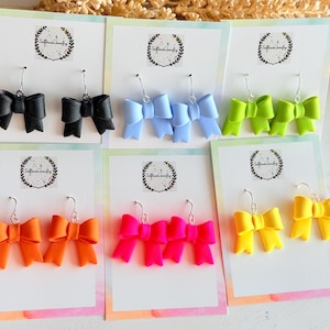 May include: A collection of handmade bow earrings in various colors, including black, light blue, green, orange, hot pink, and yellow. Each pair is displayed on a white card with a silver hook. The earrings are a fun and colorful accessory.