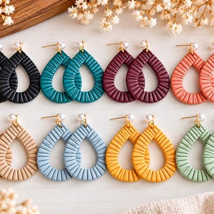 May include: Eight pairs of teardrop-shaped earrings arranged in two rows. Each earring features a textured, ribbed surface and a small white bead at the top. The colors include black, teal, dark red, coral, beige, light blue, yellow, and sage green.