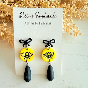 May include: Handmade earrings with a yellow disc, black bee design, black teardrop, and black bow. The earrings are displayed on a white card with the text "Blooms Handmade Selfmade by Margi".