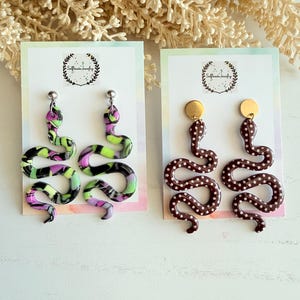 May include: Two pairs of snake-shaped dangle earrings on display cards. The left pair features a marbled pattern of neon green, black, and purple. The right pair is solid dark brown with white polka dots and gold-toned circular stud tops.