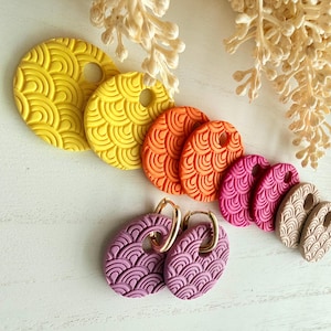 Puede incluir: Una colección de pendientes y colgantes coloridos en tonos amarillo, naranja, rosa y beige. Los pendientes tienen aros dorados. Los colgantes son redondos y ovalados con un diseño texturizado. Medidas métricas.