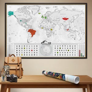 Può includere: Una mappa del mondo incorniciata con bordo nero, sfondo bianco e punti salienti dei paesi colorati. Sotto la mappa ci sono icone e un logo della mappa del mondo. Uno zaino marrone, una macchina fotografica e un poster arrotolato su un tavolo di legno.