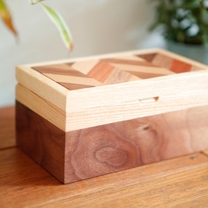 Op de afbeelding: Een houten doos met een deksel met een chevron-patroon. De doos is gemaakt van licht en donker hout en heeft een natuurlijke afwerking.