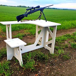 Op de afbeelding: Een witte houten schietbank met een geïntegreerde zitting en een uitgesneden ontwerp op de poten. Een zwart grendelgeweer met een gemonteerde richtkijker en een bipod staat op het platte oppervlak van de bank, klaar voor gebruik buiten.