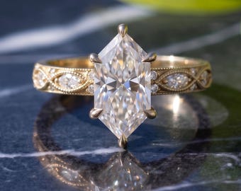 Op de afbeelding: Een gouden ring met een centrale diamant in marquise-slijpvorm, vastgehouden door zes pootjes. De band heeft een ingewikkeld gegraveerd patroon met kleine, ingelegde marquisevormige diamanten aan de zijkanten en een gepolijste afwerking.