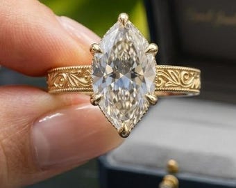 May include: A gold ring featuring a large marquise-cut center stone held by prongs. The band is crafted with an intricate, raised floral or scrollwork engraving that extends along the sides. The ring is positioned in front of a dark blue box.