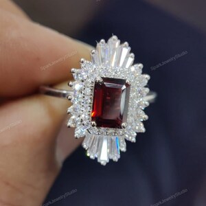 Può includere: Un anello d'argento con una grande pietra preziosa rettangolare rossa circondata da pietre preziose più piccole e trasparenti. L'anello ha un design delicato e intricato.