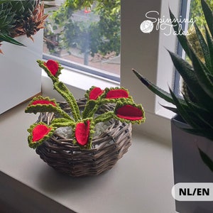 May include: A crocheted Venus flytrap plant with green stems and vibrant red interior traps, potted in a small, round, woven brown wicker basket filled with white stones. The text Spinning Tales is visible in the upper right corner.