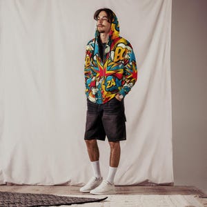 Puede incluir: Una chaqueta con capucha y cremallera con un estampado de c&oacute;mic colorido que incluye palabras como "BOOM" y "ZA". La chaqueta se lleva sobre una camiseta negra y pantalones cortos negros.