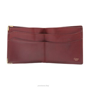 May include: A burgundy leather wallet with gold accents. The wallet is open and shows the interior with multiple card slots and a bill compartment. The wallet has the Cartier Paris logo on the bottom right corner.