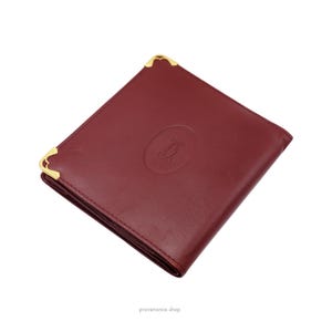 May include: A burgundy leather wallet with gold accents and a Cartier logo embossed on the front.