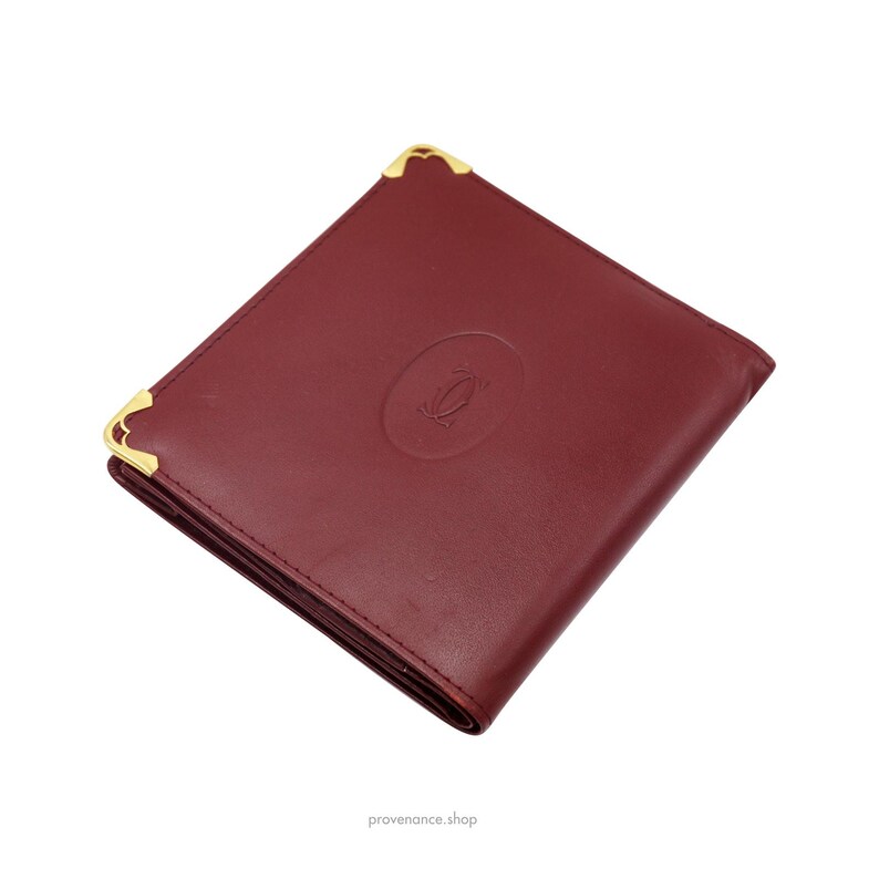 May include: A burgundy leather wallet with gold accents and a Cartier logo embossed on the front.