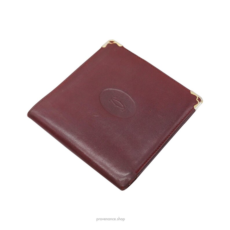 May include: A burgundy leather wallet with gold corners and a gold embossed Cartier logo.