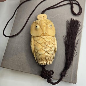 May include: A cream-colored carved owl pendant featuring a smaller owl figure on its head. The owl has detailed feather textures and dark eyes. It is attached to a dark brown cord with a decorative knot and a long, thin matching tassel.