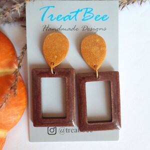 Op de afbeelding: Een paar bruine en gouden statement oorbellen met een rechthoekig frame en een traanvormige hanger. De oorbellen zijn handgemaakt en hebben een glinsterende gouden afwerking.