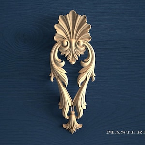 May include: A light-colored wooden decorative carving featuring intricate acanthus leaf scrollwork and a central fan-shaped crest. The symmetrical ornament displays detailed relief patterns and a small tassel-like element at the bottom point.