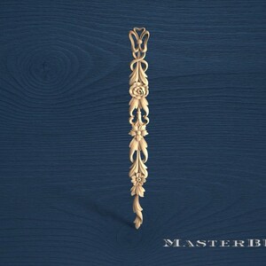 May include: A carved wooden decorative element featuring floral and leaf motifs. The design includes a rose, stylized leaves, and a bow-like top. The piece is light brown against a dark blue wood grain background.