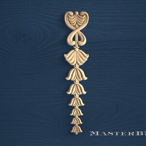 May include: A carved wooden decorative element with a repeating floral design. The piece is light brown and has a symmetrical shape, with a series of stylized flower-like forms. The background is a dark blue wood grain.