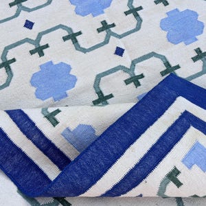 May include: A white rug with a blue and green geometric pattern. The rug has a blue border with white stripes.