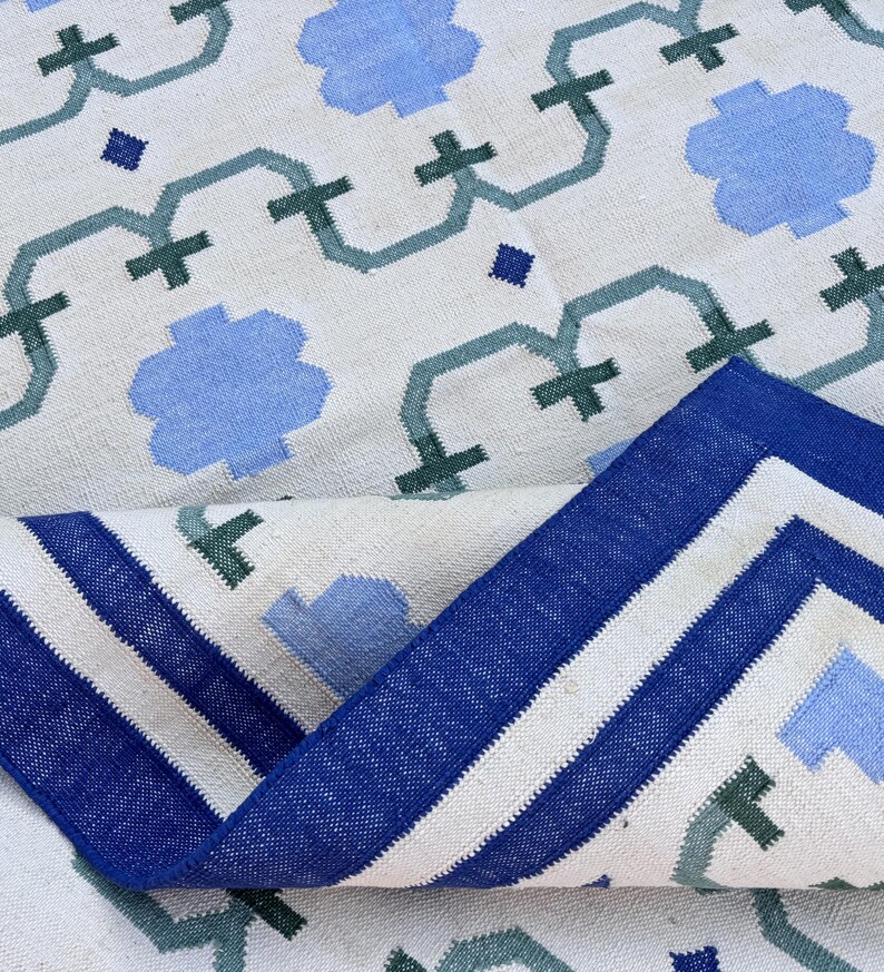 May include: A white rug with a blue and green geometric pattern. The rug has a blue border with white stripes.