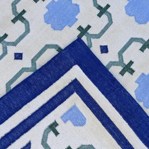 May include: A white rug with a blue and green geometric pattern. The rug has a blue border with a white stripe.