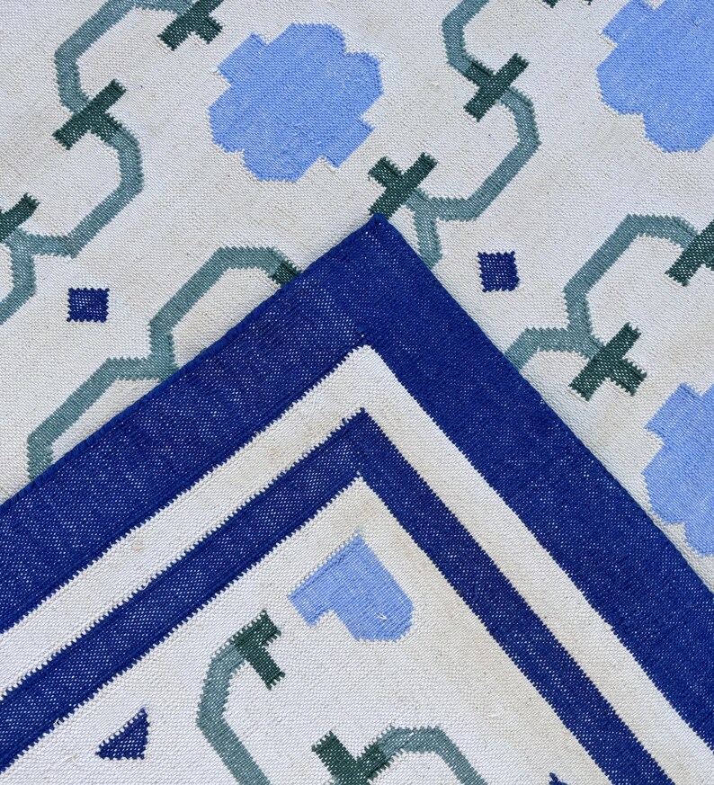 May include: A white rug with a blue and green geometric pattern. The rug has a blue border with a white stripe.