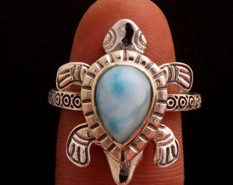 May include: A silver ring featuring a turtle design. The ring has a light blue, teardrop-shaped stone at the center. The turtle's shell and flippers are detailed with etched patterns. The ring is displayed against a dark background.